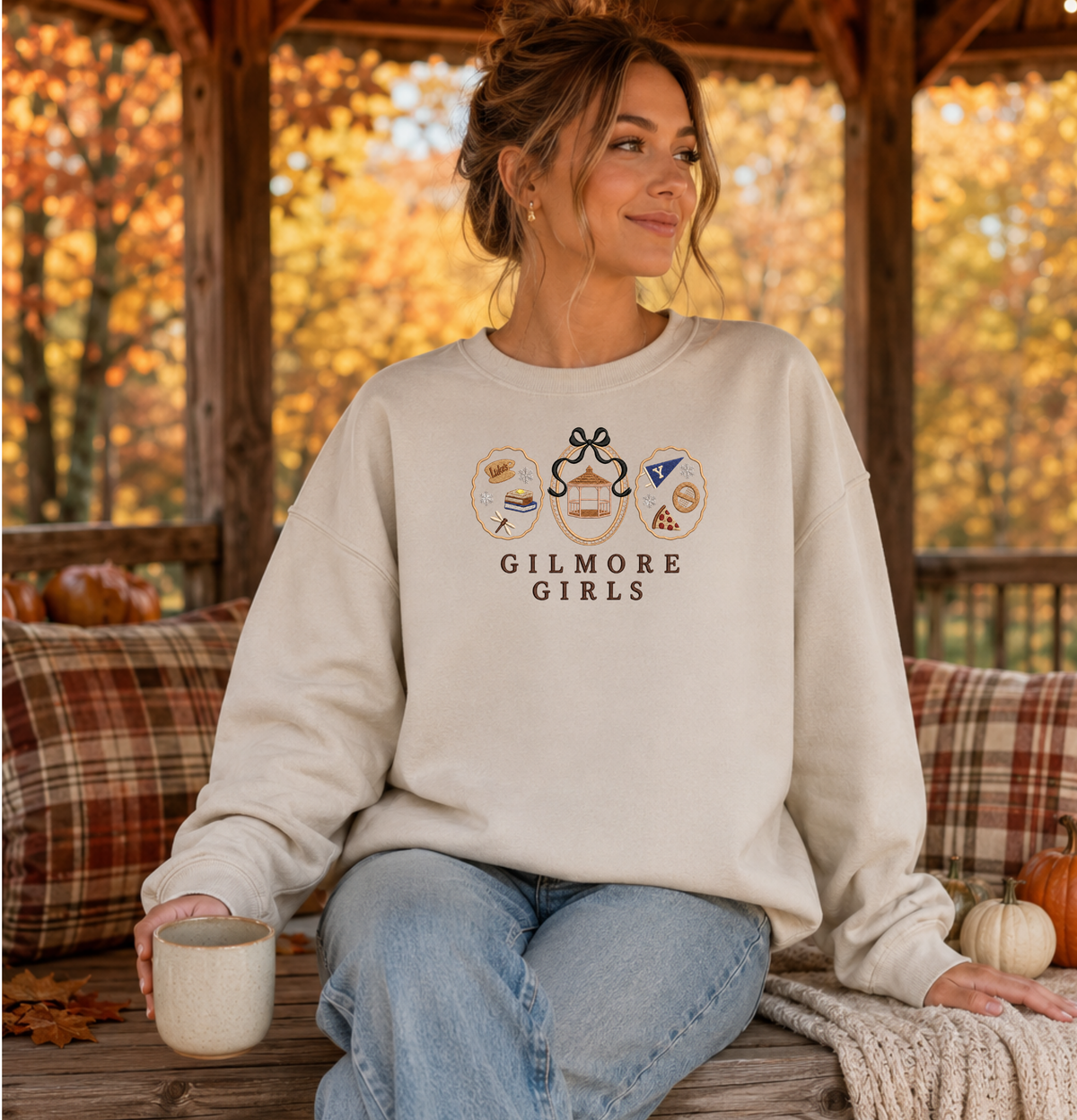 Woman wearing a sand Gilmore Girls embroidered crewneck sweatshirt featuring Stars Hollow icons and a black bow.