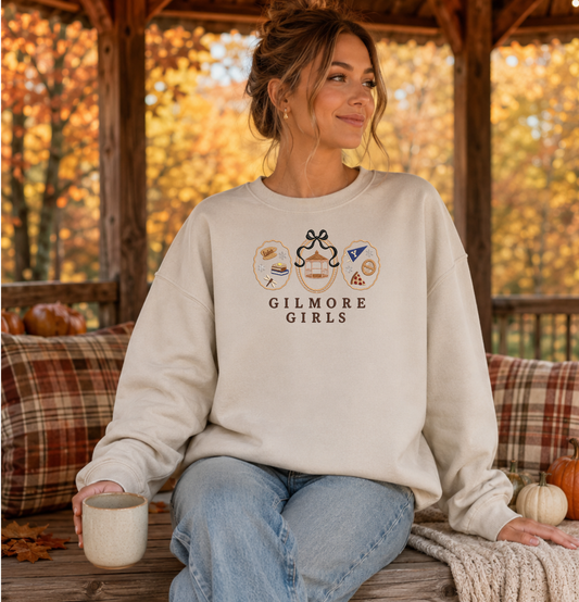 Woman wearing a sand Gilmore Girls embroidered crewneck sweatshirt featuring Stars Hollow icons and a black bow.